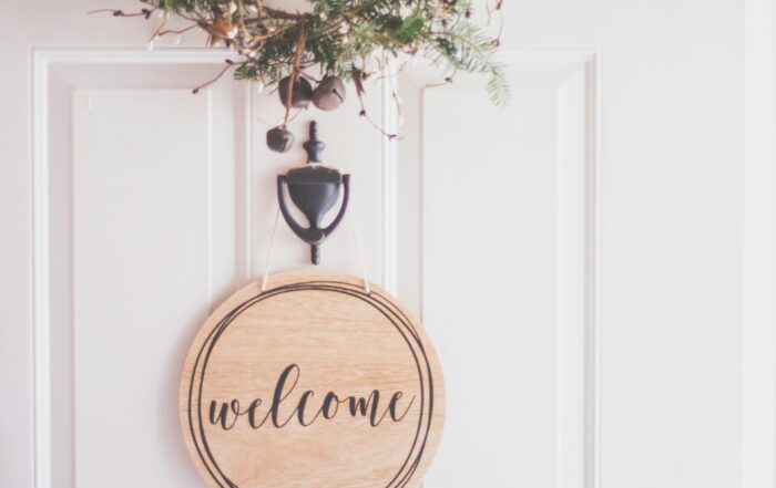 Pintu berwarna putih dengan papan bertuliskan welcome dan hiasan daun dan lonceng