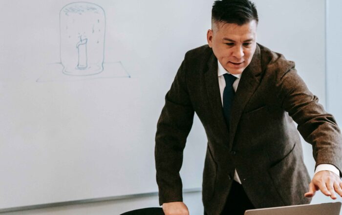 Pria mengenakan jas dengan senyum tipis duduk di depan laptop, naskah MC webinar online