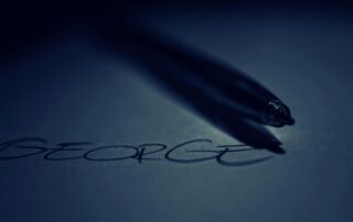 sebuah pena yang sedang menuliskan nama "George" di atas kertas. Pena tersebut menghasilkan bayangan yang panjang dan tipis di atas kertas, seakan-akan sedang menari di atas kata yang baru saja ditulis.