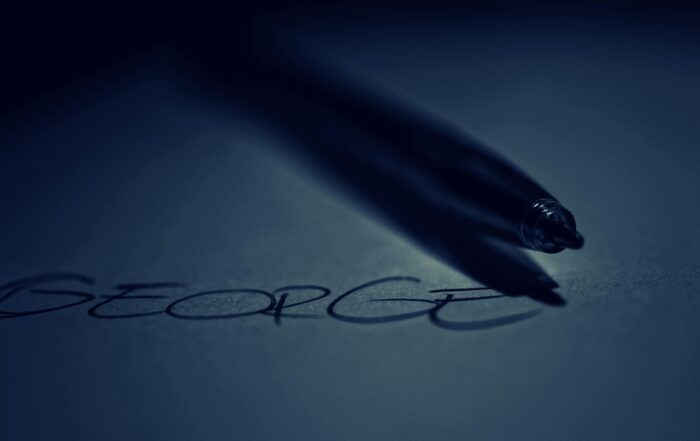 sebuah pena yang sedang menuliskan nama "George" di atas kertas. Pena tersebut menghasilkan bayangan yang panjang dan tipis di atas kertas, seakan-akan sedang menari di atas kata yang baru saja ditulis.