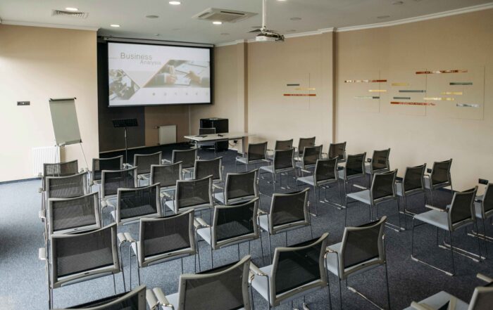 Auditorium dengan meja dan kursi tersusun rapi. Terdapat layar proyektor untuk menampilkan bahan presentasi