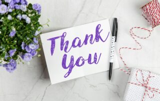 Kartu bertuliskan terima kasih dengan seikat bunga ungu di sampingnya. Terdapat pena dan kotak hadiah dengan latar belakang putih