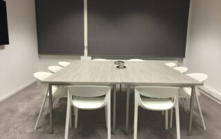 Ruang rapat dengan meja persegi yang besar dan beberapa kursi berwarna putih. tampak karpet berwarna abu dan tirai putih untuk masuknya cahaya. Di atas meja terdapat alat tulis untuk rapat. Jadi MC andal