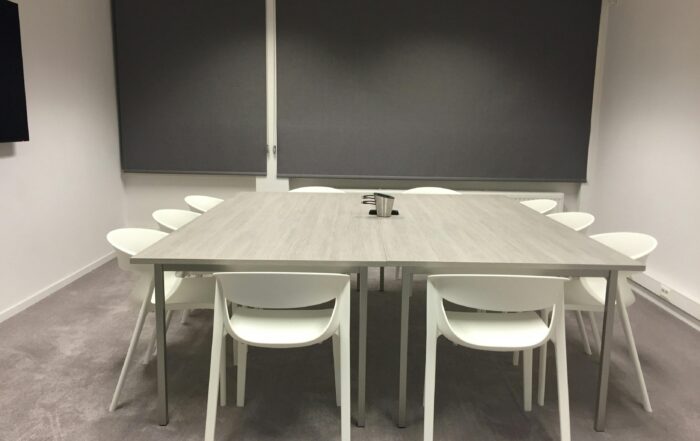 Ruang rapat dengan meja persegi yang besar dan beberapa kursi berwarna putih. tampak karpet berwarna abu dan tirai putih untuk masuknya cahaya. Di atas meja terdapat alat tulis untuk rapat. Jadi MC andal