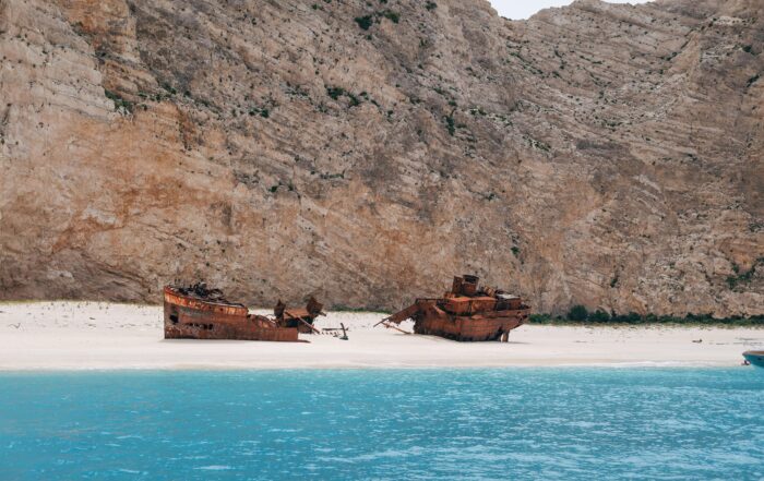Kapal karam yang sudah berkarat di atas pasir putih dengan latar belakang pantai yang berwarnaa biru dan tebing akrang tinggi hari disabilitas internasional
