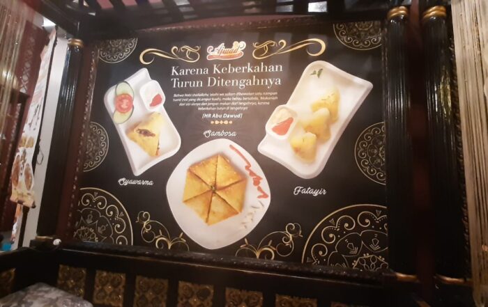 poster menu makanan di sebuah restoran. Poster ini didominasi oleh warna gelap dengan tulisan berwarna emas yang mencolok, memberikan kesan mewah dan elegan. Menu makanan di restoran Timur Tengah di Jakarta yang tampak adalah kubba, samosa, dan fatayir.