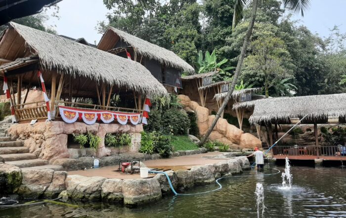 pemandangan yang asri dan alami. Terdapat beberapa bangunan dengan atap jerami yang khas rumah tradisional Indonesia, dibangun di atas sebuah area berundak dengan kolam air di bagian bawahnya. Beberapa pohon palem tumbuh di sekitar area tersebut, menambah kesan hijau dan sejuk.